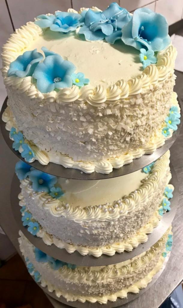Trzypiętrowy tort weselny z białym kremem, posypką z białej czekolady i dekoracjami z błękitnych kwiatów, umieszczony na metalowych paterach.