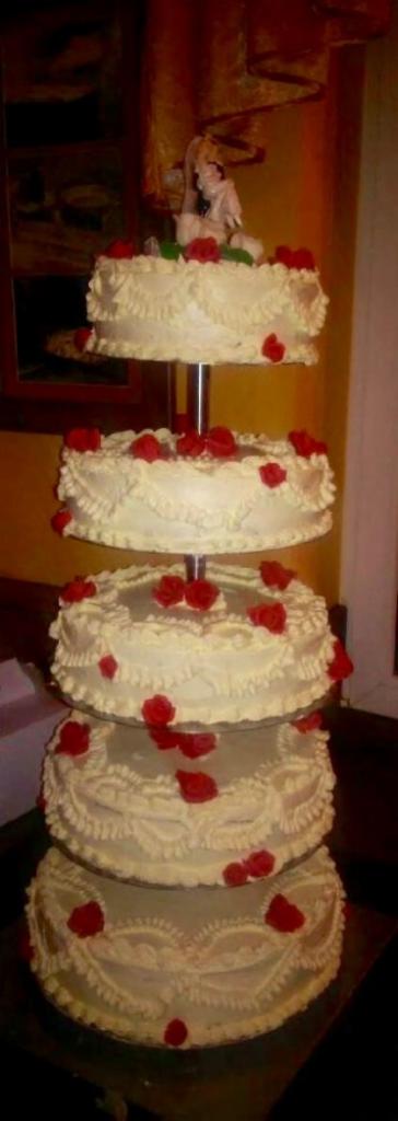Czteropiętrowy tort weselny w kolorze kremowym, zdobiony czerwonymi różami i figurką pary młodej na szczycie, ustawiony na metalowym stelażu.