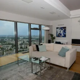 Jasne wnętrze apartamentu z panoramicznym widokiem na miasto, beżowa sofa z poduszkami, szklany stolik kawowy na dywanie, nowoczesne oświetlenie i abstrakcyjny obraz na ścianie.