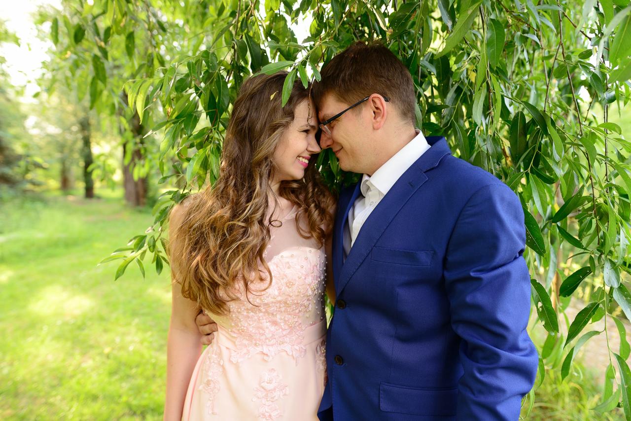 Uśmiechnięta para w objęciach, panna młoda w pudrowo-różowej sukni z koronkowymi aplikacjami, pan młody w niebieskim garniturze, stoją pod drzewem w słoneczny dzień.