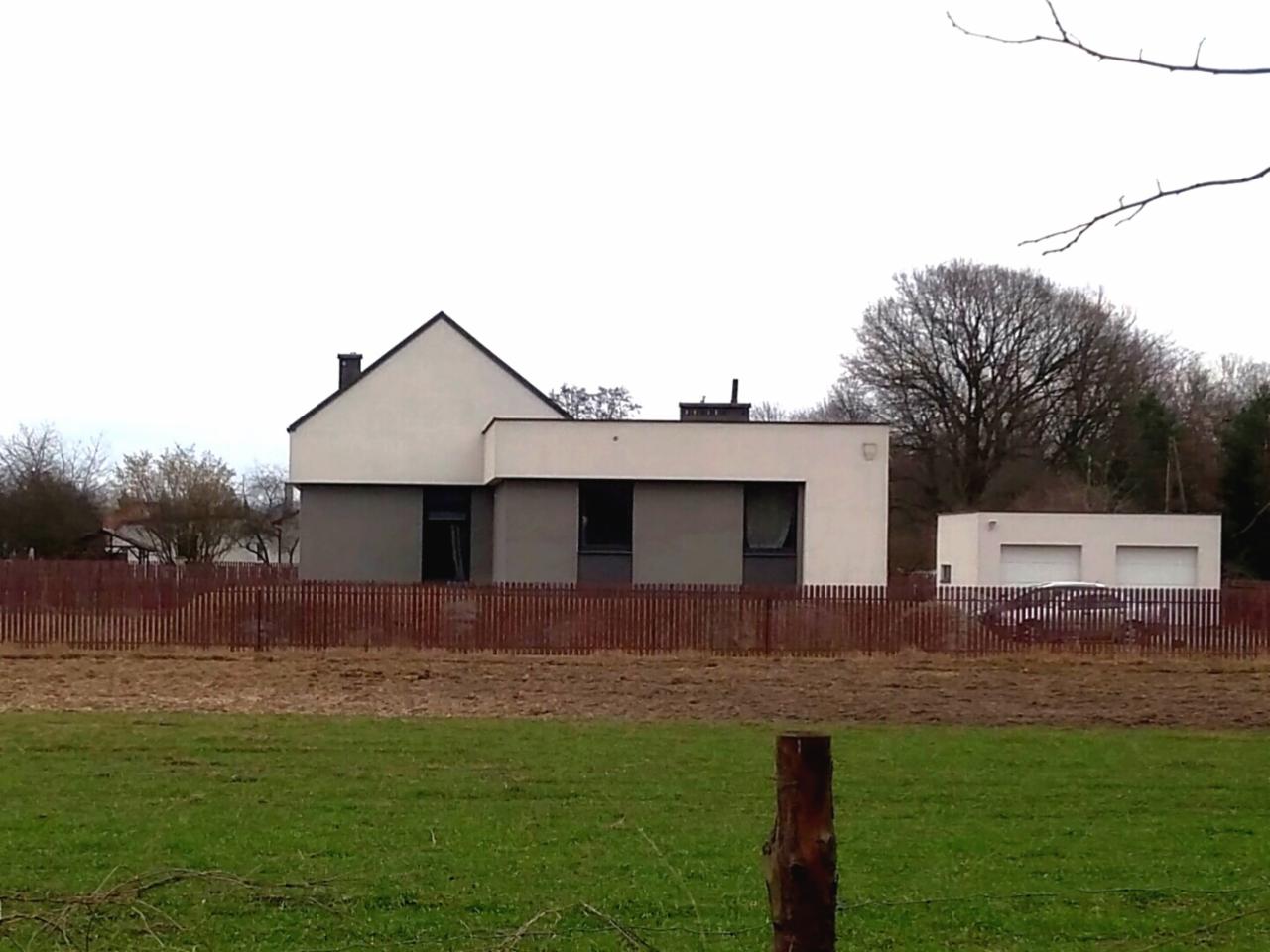 Nowoczesny dom jednorodzinny z płaskim dachem i elewacją w odcieniach bieli i szarości, otoczony drewnianym płotem i zielonym trawnikiem, widoczny w pochmurny dzień.