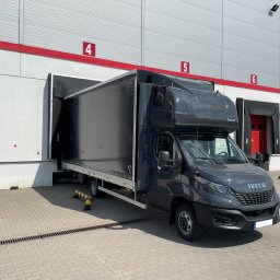Przedsiębiorstwo Handlowo-Usługowe Piotr Tuszyński - Czarny samochód dostawczy Iveco z zabudową skrzyniową, częściowo w doku magazynowym z numerem 4, na tle szaro-białej elewacji. Widok z przodu, słoneczny dzień.
