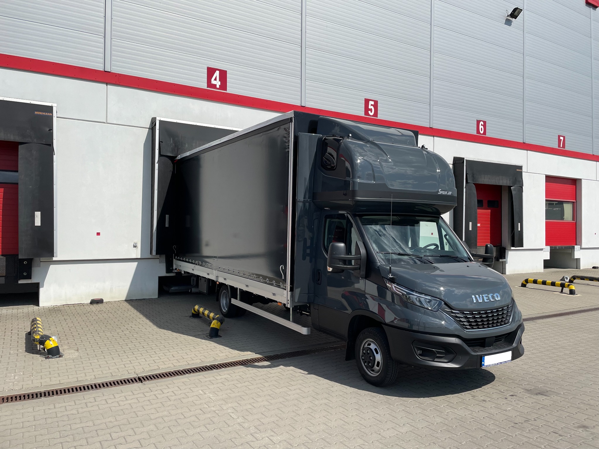 Czarny samochód dostawczy Iveco z zabudową skrzyniową, częściowo w doku magazynowym z numerem 4, na tle szaro-białej elewacji. Widok z przodu, słoneczny dzień.