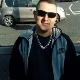 Mężczyzna w okularach przeciwsłonecznych i bluzie z kapturem, pozujący na tle ulicy z samochodami i budynkami mieszkalnymi.