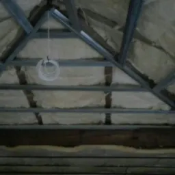 Skośny sufit w trakcie ocieplania wełną mineralną, widoczne metalowe profile konstrukcyjne i zwisający kabel elektryczny.