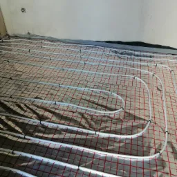 Białe rury ogrzewania podłogowego ułożone na folii z czerwoną siatką na surowym podłożu, widoczne fragmenty ścian w stanie deweloperskim.