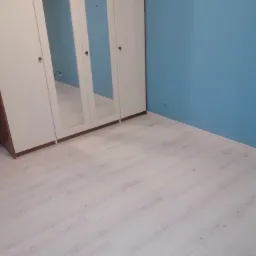 Jasne panele podłogowe w pokoju z błękitną ścianą i białą szafą z lustrami.