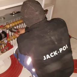 Pracownik firmy JACK-POL w czarnej bluzie i czapce, w trakcie montażu rozdzielacza ogrzewania podłogowego w białej szafce, używając zaciskarki do rur PEX; widoczne czerwone rury na podłodze.