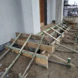 Szczegółowy widok szalunku fundamentu przy ścianie budynku w trakcie budowy, z widocznym zbrojeniem i ziemią wewnątrz, oraz stertą odpadów budowlanych w tle.