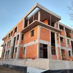 Wielopiętrowy budynek w trakcie budowy z czerwonej cegły i betonu, widoczne podpory stropu na ostatniej kondygnacji, błękitne niebo w tle.