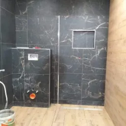 Łazienka w trakcie wykańczania: ciemne płytki imitujące marmur na ścianach, drewnopodobne płytki na podłodze, widoczne elementy instalacji sanitarnej, w tle ściana wyłożona drewnianą boazerią.