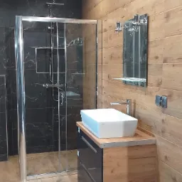 Nowoczesna łazienka z kabiną prysznicową typu walk-in, umywalką nablatową na szafce z szufladami i lustrem z wieszakami na ręczniki, wykończona płytkami imitującymi drewno i czarnym marmurem.