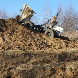 Operator w pomarańczowym kombinezonie obsługuje wywrotkę na zboczu wykopu, wysypując ziemię. Widoczne warstwy gleby w przekroju.