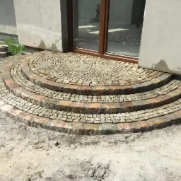 Półokrągłe schody wejściowe ułożone z kostki granitowej w odcieniach szarości i brązu, prowadzące do przeszklonych drzwi budynku w trakcie budowy lub remontu.