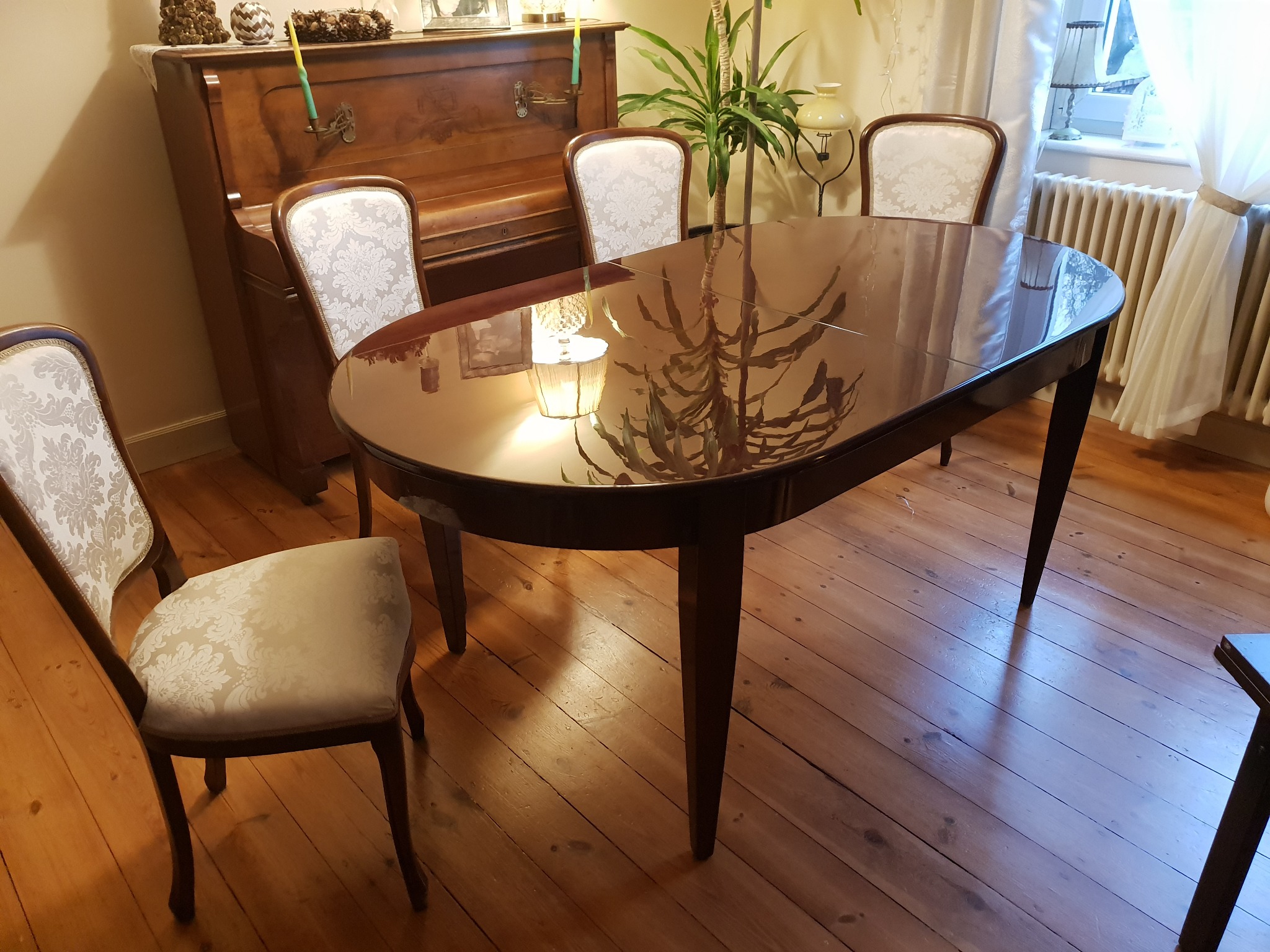 Rozkładany stół z ciemnego drewna o owalnym blacie, otoczony krzesłami z tapicerką w kwiatowy wzór, w tle pianino i roślina doniczkowa.