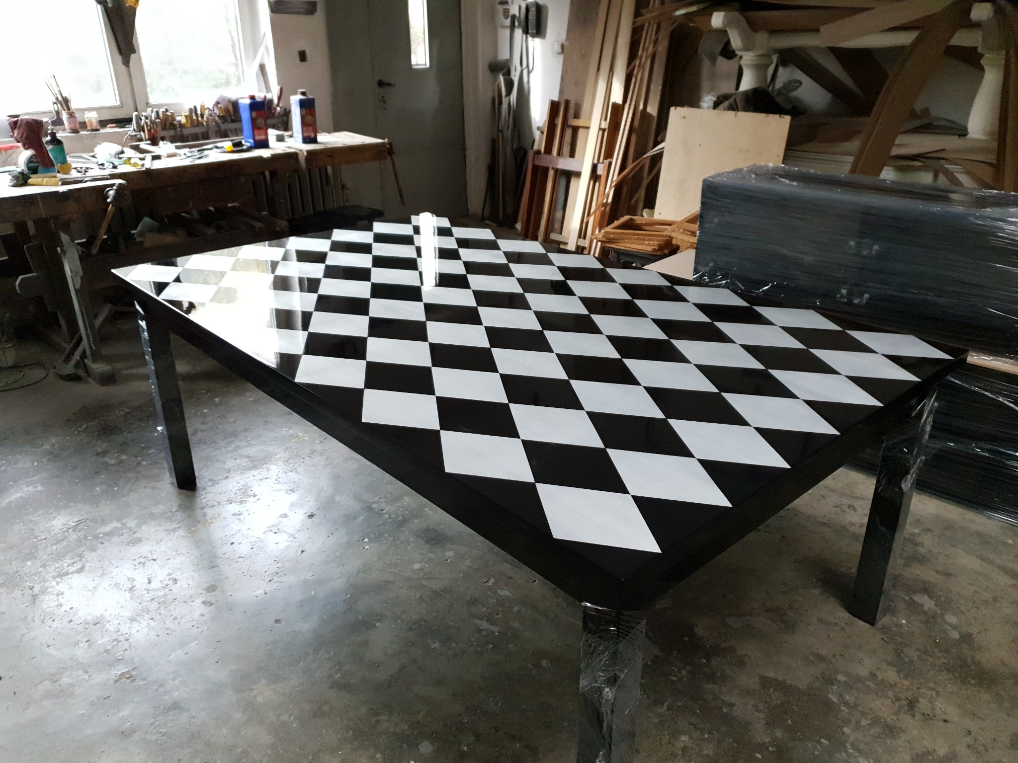 Lakierowany stół z blatem w szachownicę czarno-białą, wykonany w warsztacie stolarskim, z widocznymi narzędziami i materiałami w tle.