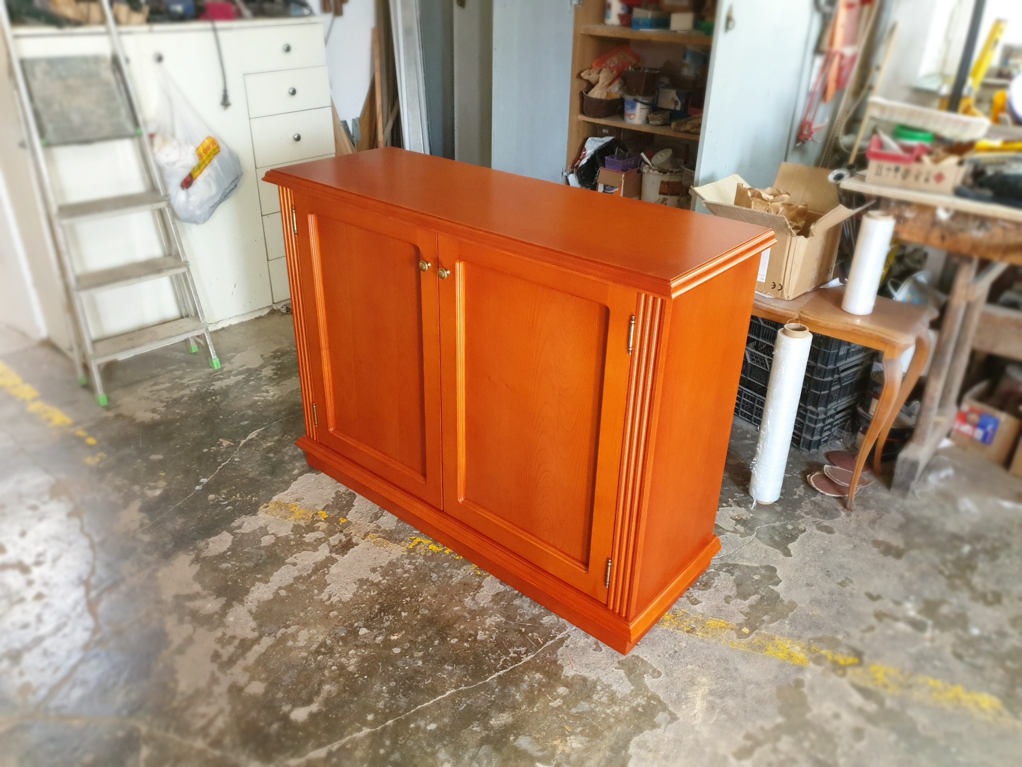 Pomarańczowa szafka z drewna z dwoma drzwiczkami i złotymi gałkami, widoczna w warsztacie na tle regałów i drabiny.