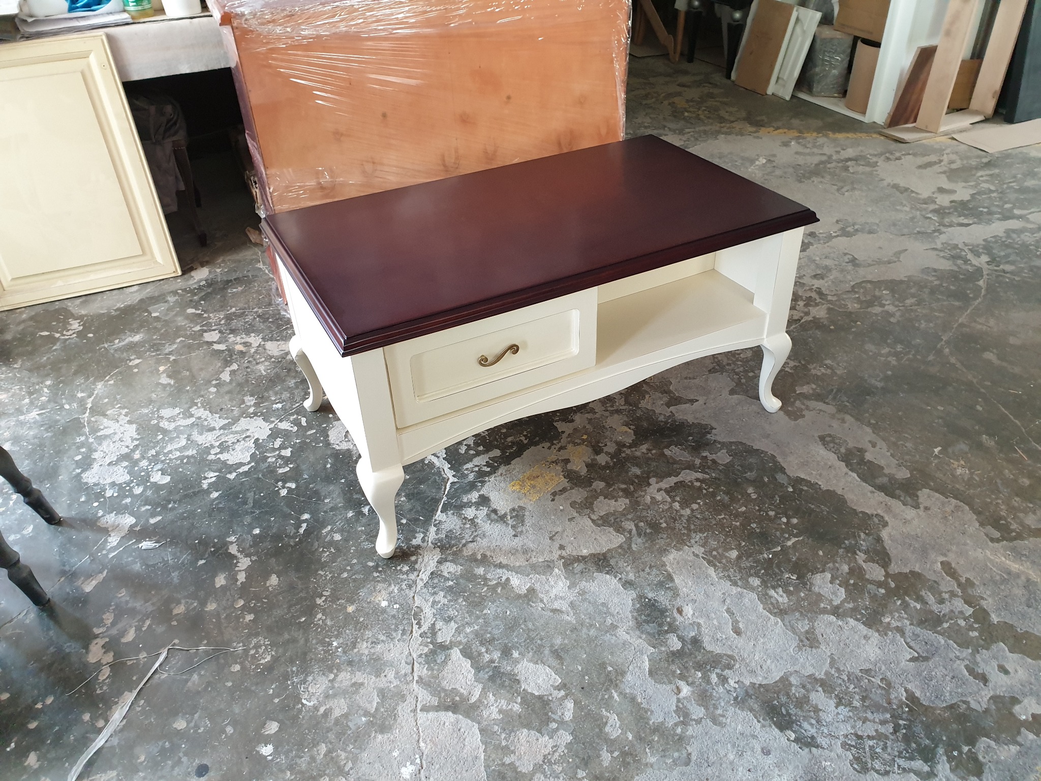 Stolik kawowy z mahoniowym blatem i kremowymi nogami z szufladą, wyprodukowany w Swarzędzu, stoi na betonowej posadzce warsztatu.