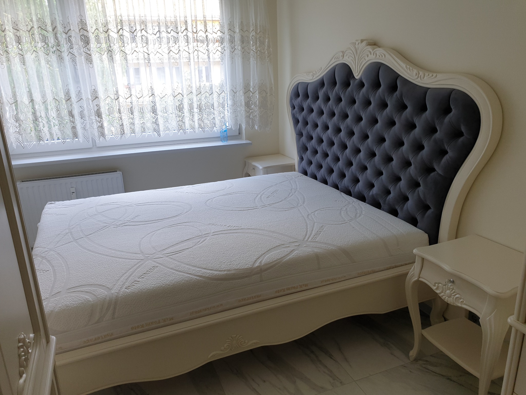 Eleganckie łóżko z pikowanym, szarym wezgłowiem i białym materacem w jasnej sypialni z oknem i koronkowymi firankami, widoczne stoliki nocne z dekoracyjnymi uchwytami.