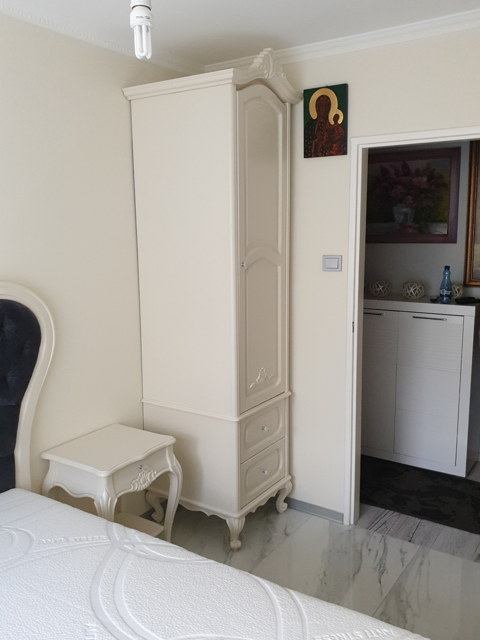 Elegancka szafa w stylu Ludwika Filipa, z ornamentami i szufladami, ustawiona w sypialni obok łóżka i stolika nocnego, z widoczną ikoną na ścianie.