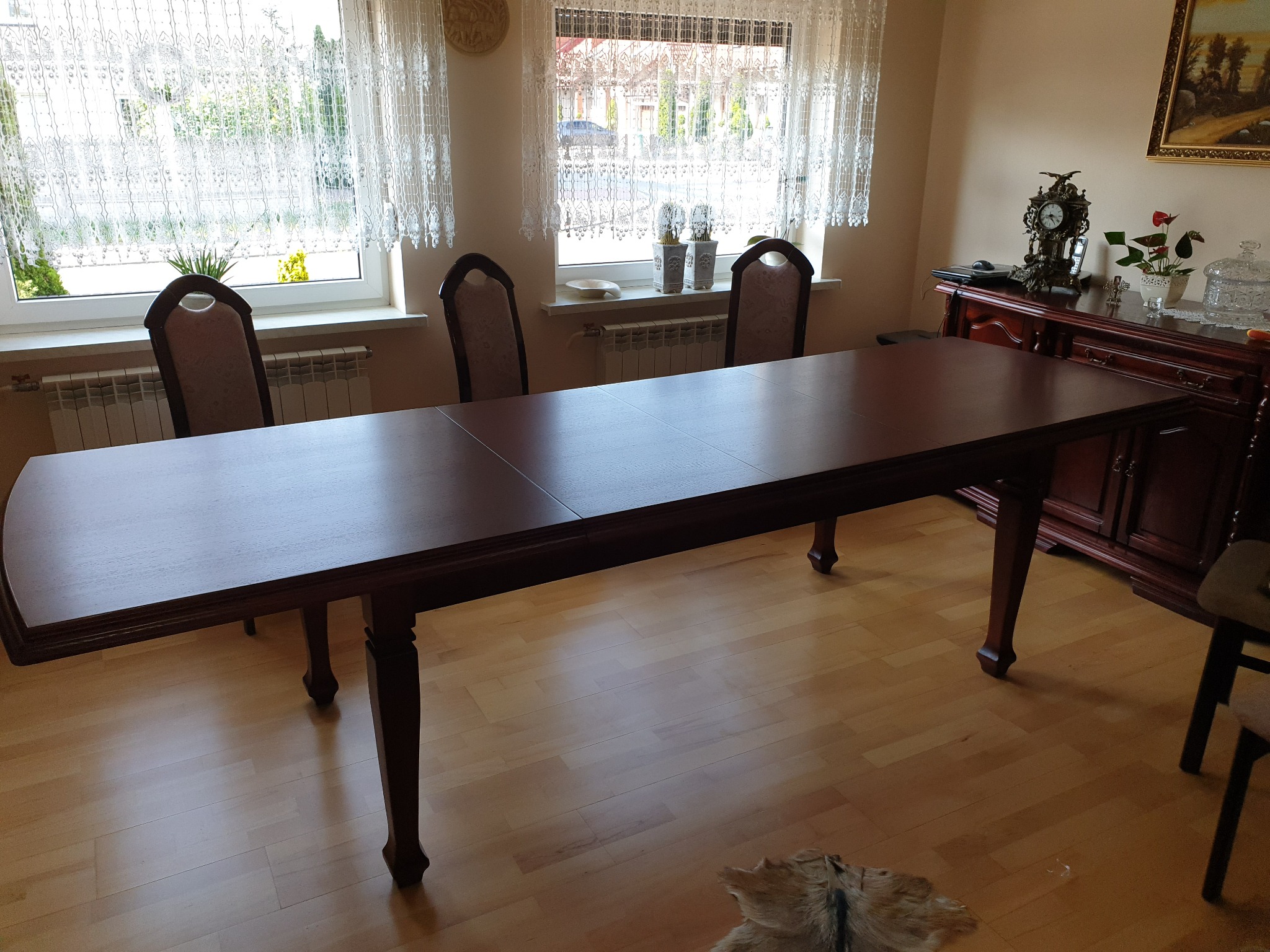 Rozkładany stół z ciemnego drewna w jadalni, z krzesłami z tapicerowanymi oparciami i widokiem na okno z koronkowymi firankami.