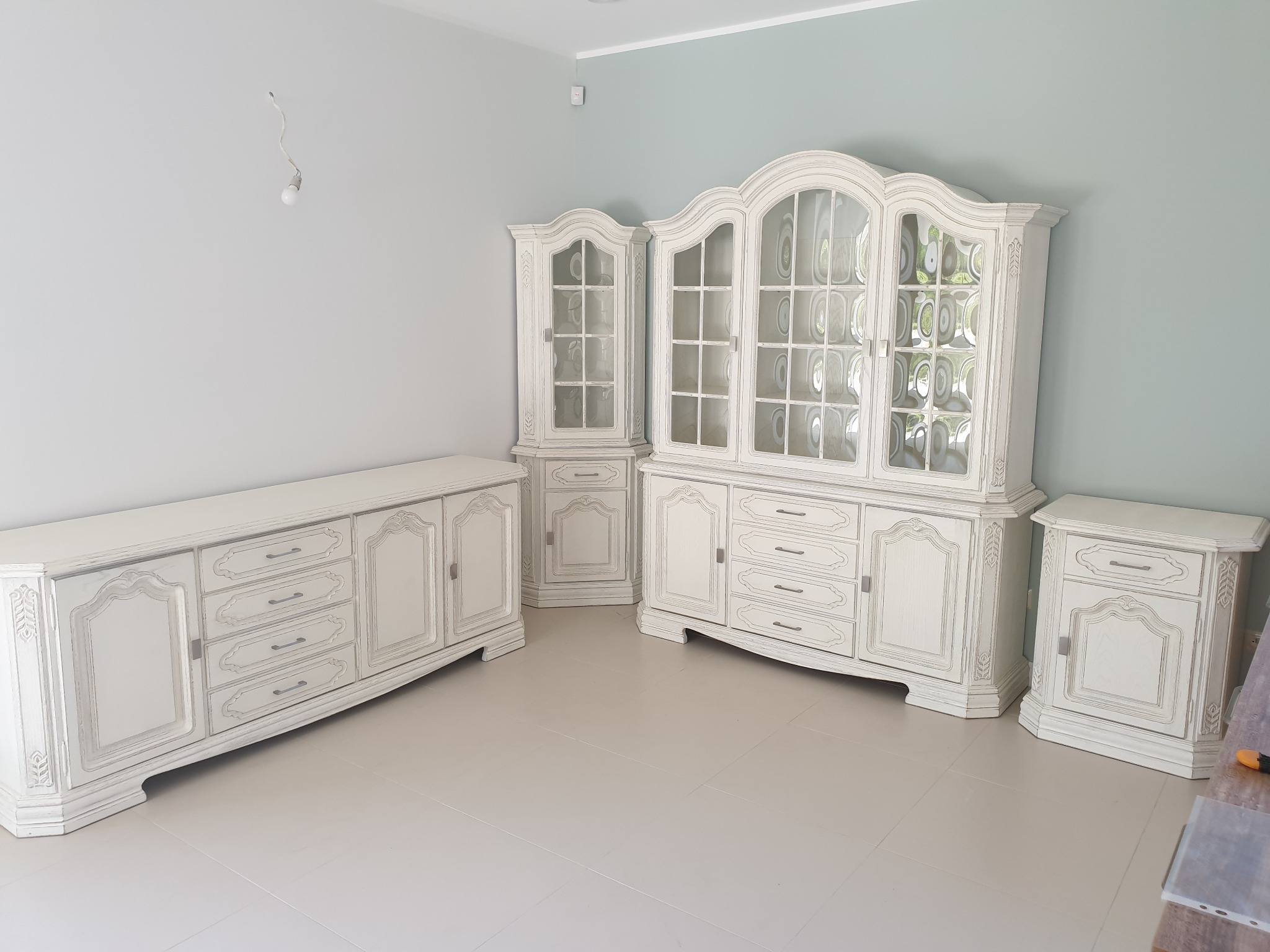 Białe, stylizowane meble drewniane w jasnym pomieszczeniu: witryna z przeszkleniami, komoda z szufladami i szafka nocna, całość w stylu vintage.