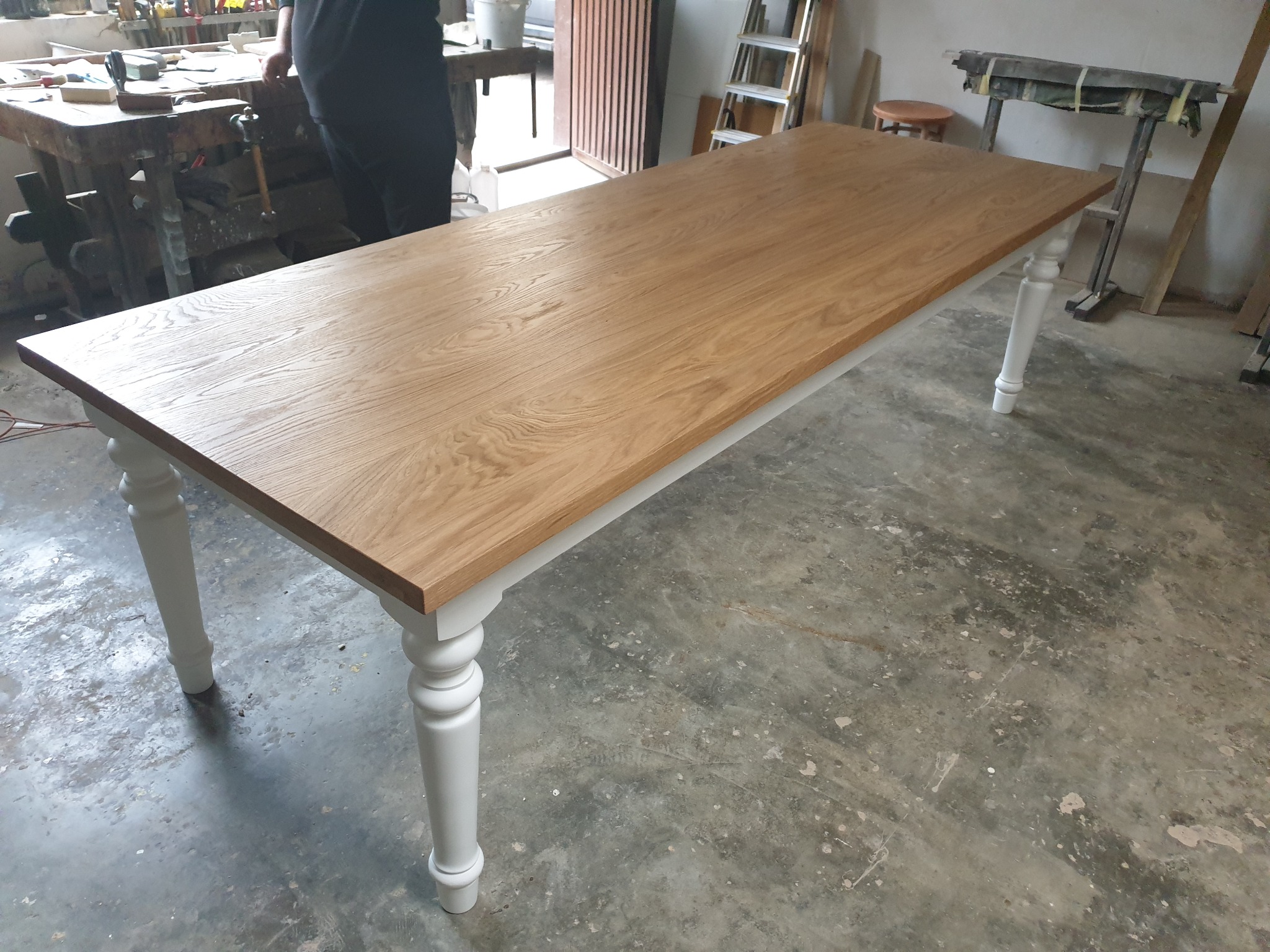 Blat dębowego stołu z białymi, toczonymi nogami, widoczny w warsztacie stolarskim z narzędziami w tle.