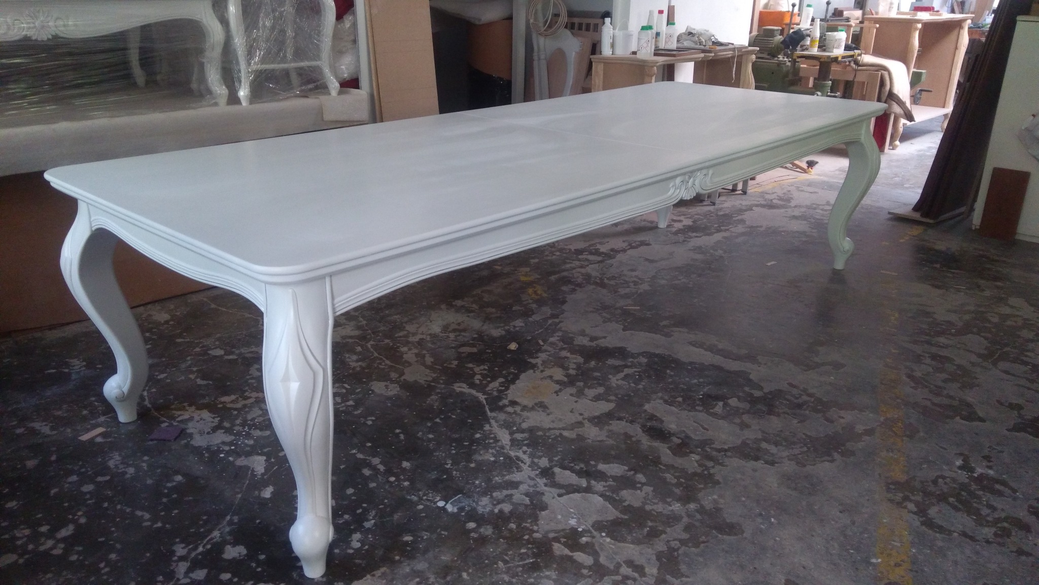 Biały, lakierowany stół w stylu Ludwika Filipa z rzeźbionymi nogami, w warsztacie stolarskim z widocznymi śladami pracy na podłodze.