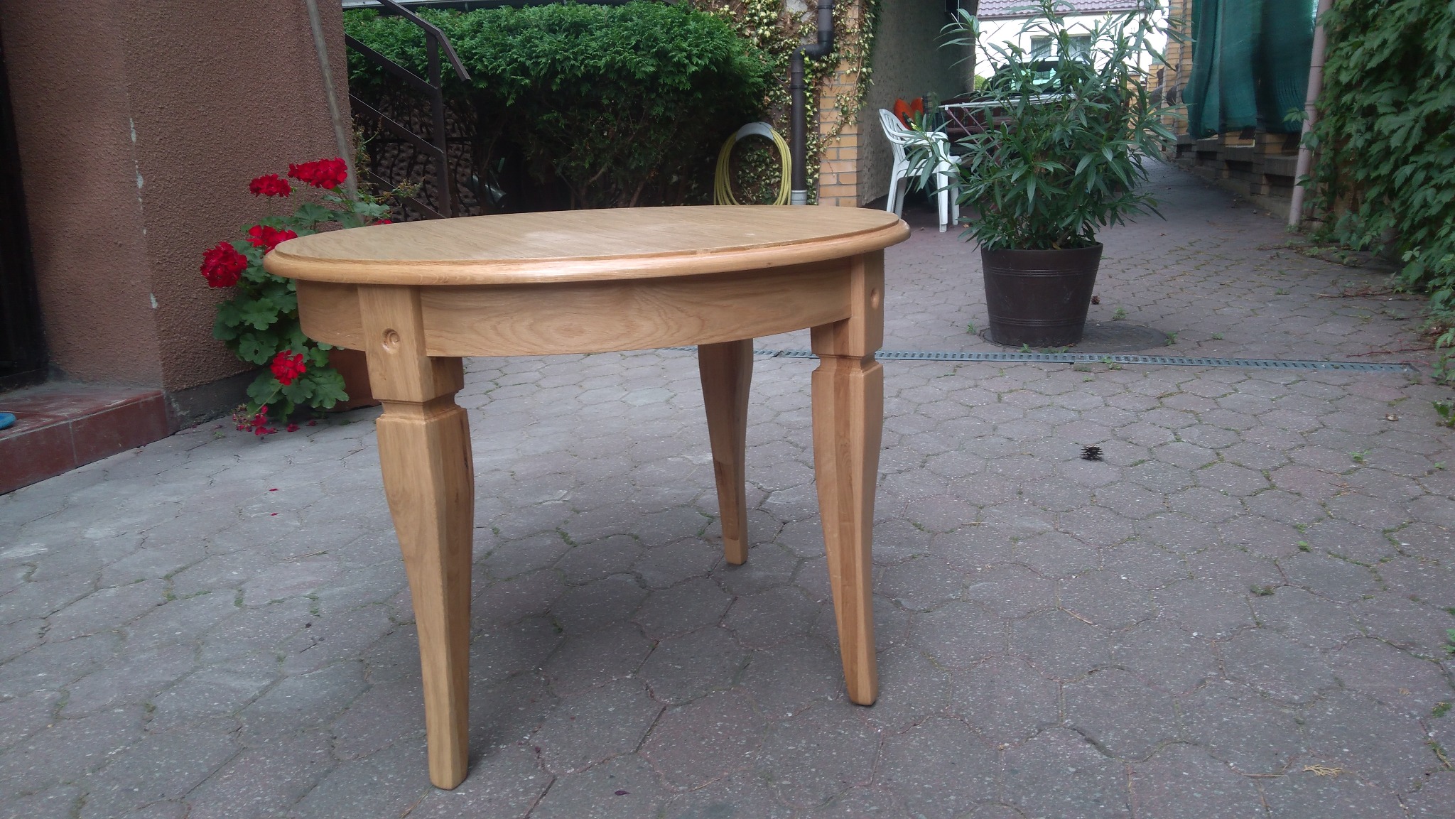 Owalny, jasny stolik z drewna ustawiony na brukowanym podwórku, z widocznymi detalami połączeń nóg, w tle donica z kwiatami i zielona roślinność.