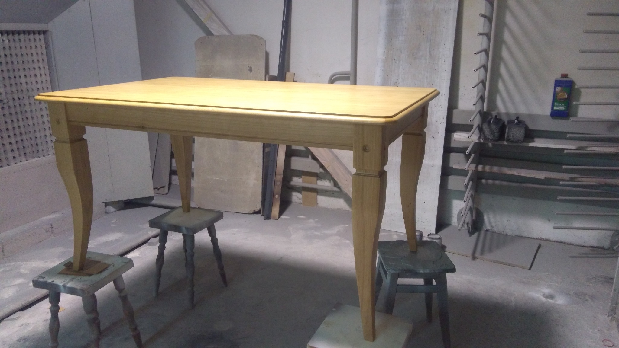 Drewniany stół na toczonych nogach, ustawiony na stołkach podczas malowania, w tle warsztat stolarski z półkami i narzędziami.