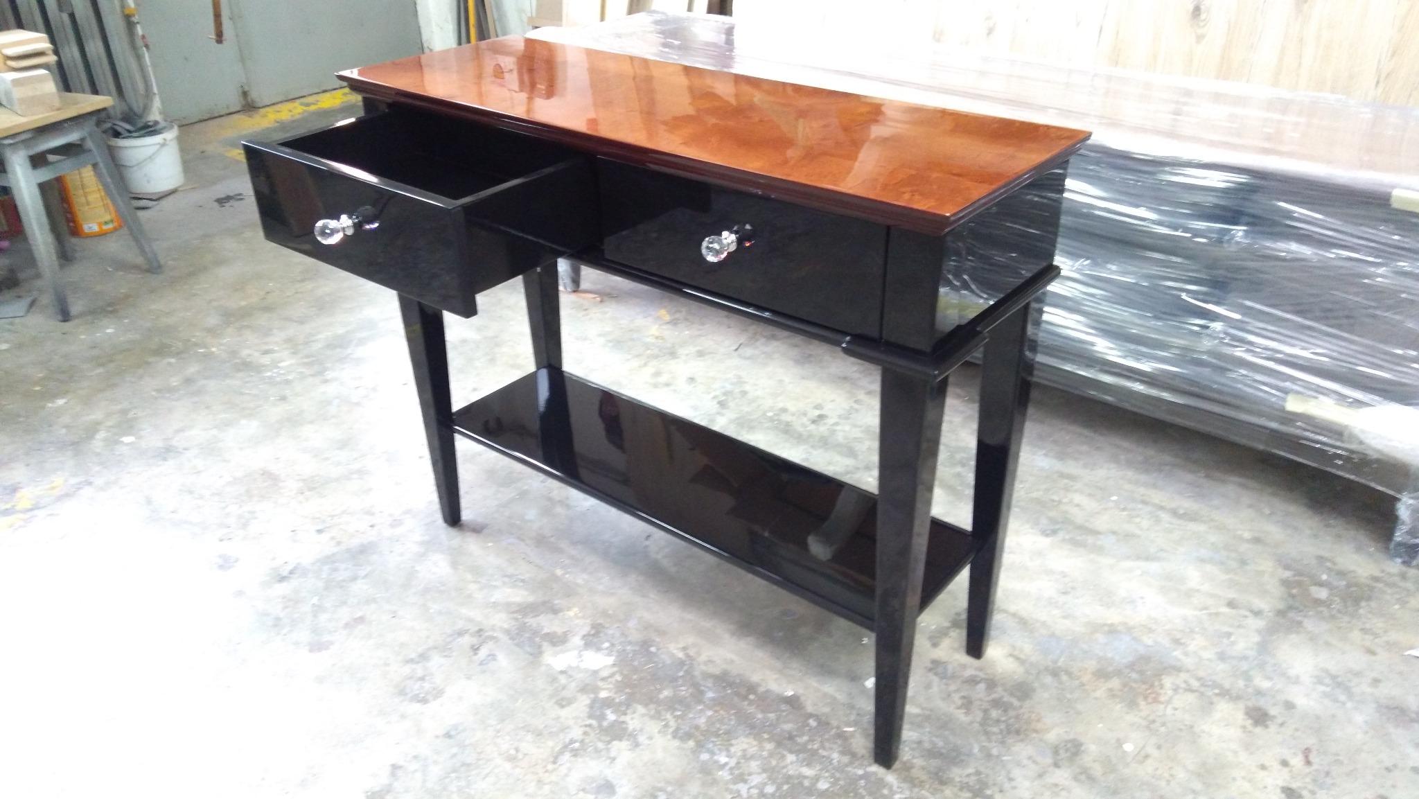 Czarna konsola z dwoma szufladami i półką, blat w kolorze orzecha, uchwyty szuflad w kształcie kryształowych gałek, na betonowej podłodze.