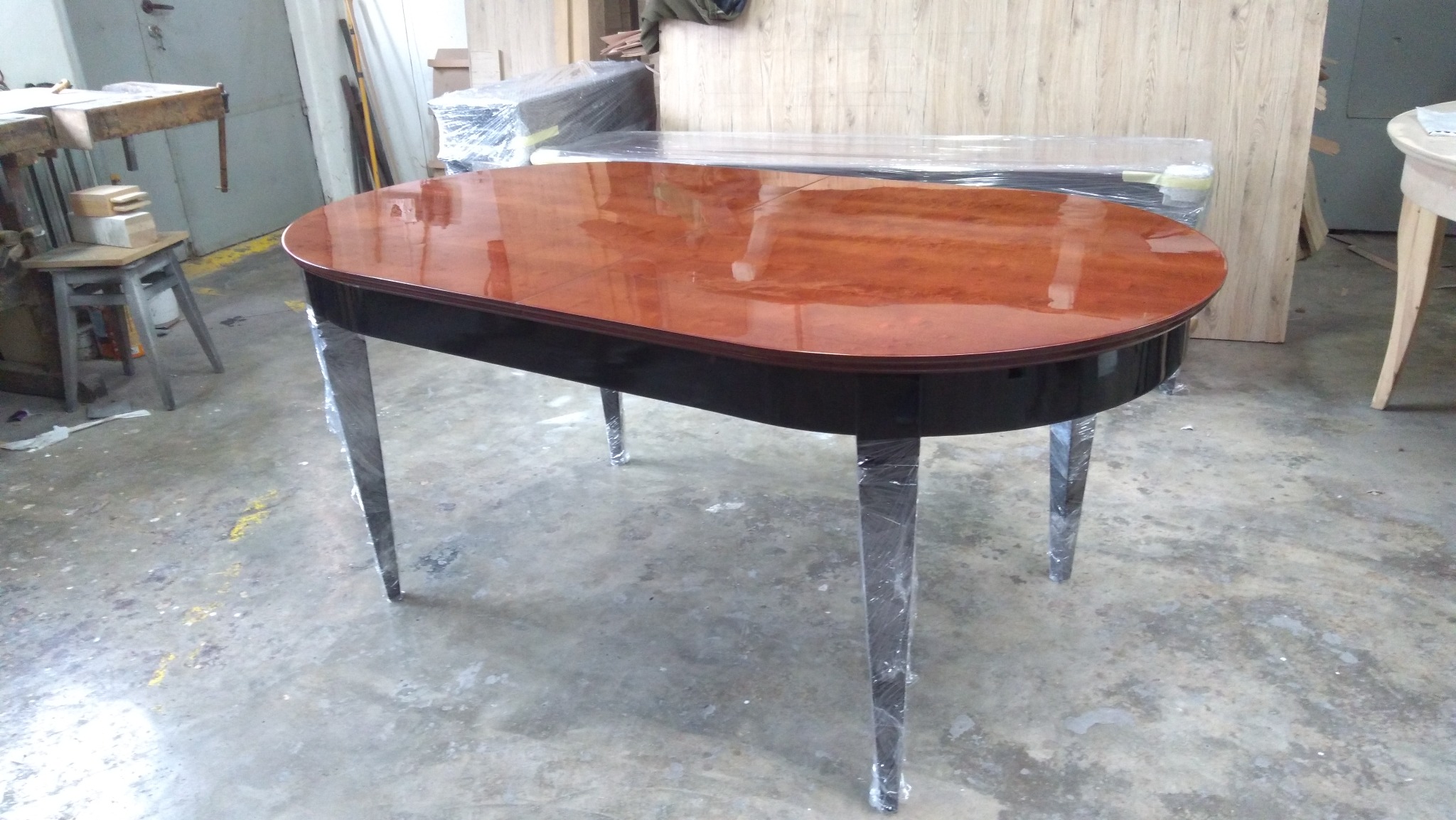 Owalny stół z lakierowanym, brązowym blatem i czarnym obrzeżem, z nogami owiniętymi folią ochronną, stojący w warsztacie.