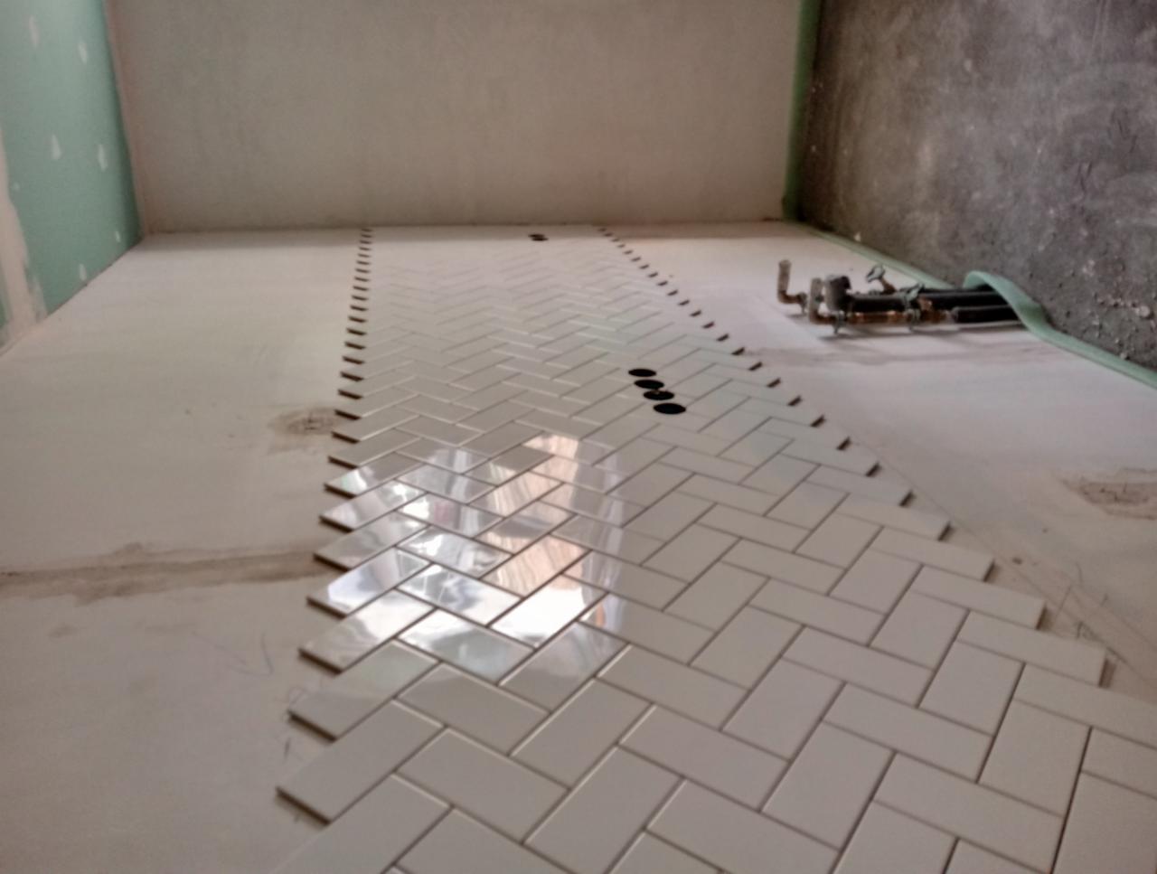 Układanie białych, błyszczących płytek w jodełkę na podłodze w trakcie prac wykończeniowych, widoczne niedokończone krawędzie i instalacja hydrauliczna przy ścianie.