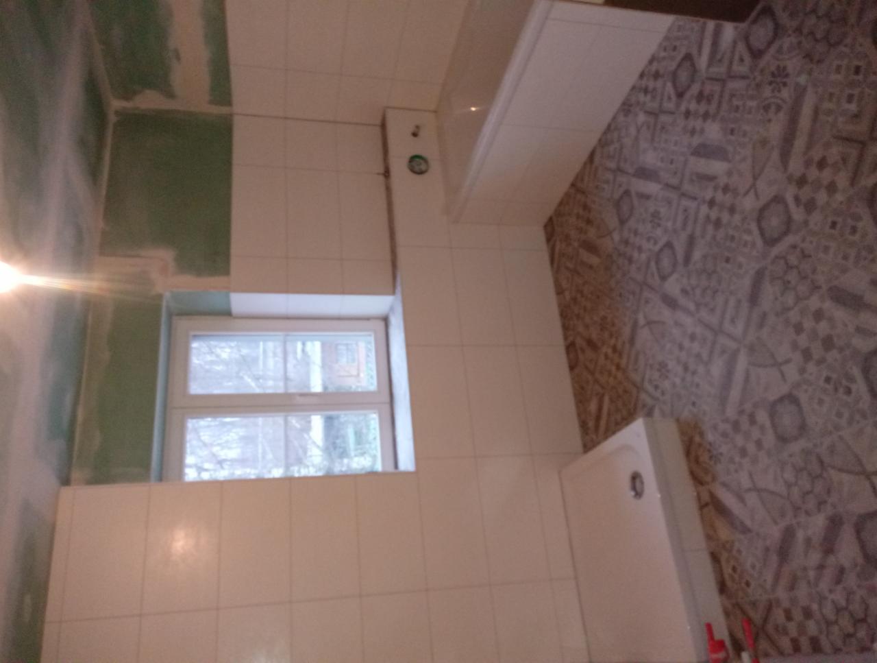 Układanie białych płytek w łazience w Chojnowie, widoczne okno, częściowo wykończona ściana z płyt gipsowo-kartonowych i wzorzysta podłoga.