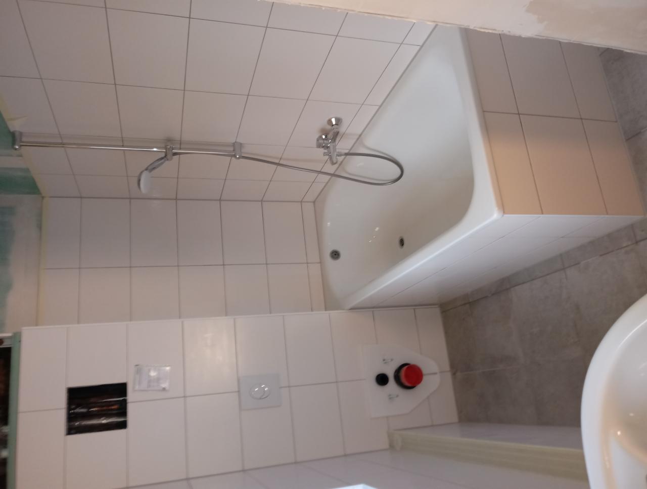 Biała wanna w obudowie z płytek, zamontowany panel prysznicowy, widoczny fragment instalacji podtynkowej, białe płytki na ścianach.