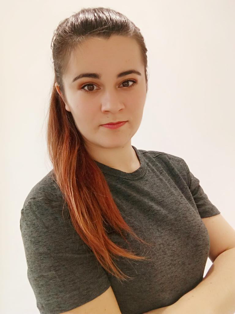 Portret kobiety z rudymi włosami spiętymi w kucyk, ubrana w szarą koszulkę, pozująca na jasnym tle.