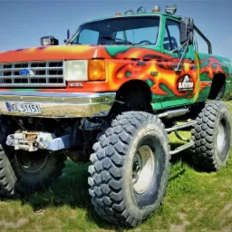 Monster truck z jaskrawym malowaniem w odcieniach zieleni, pomarańczy i żółci, stojący na trawiastym terenie z budynkiem w tle.