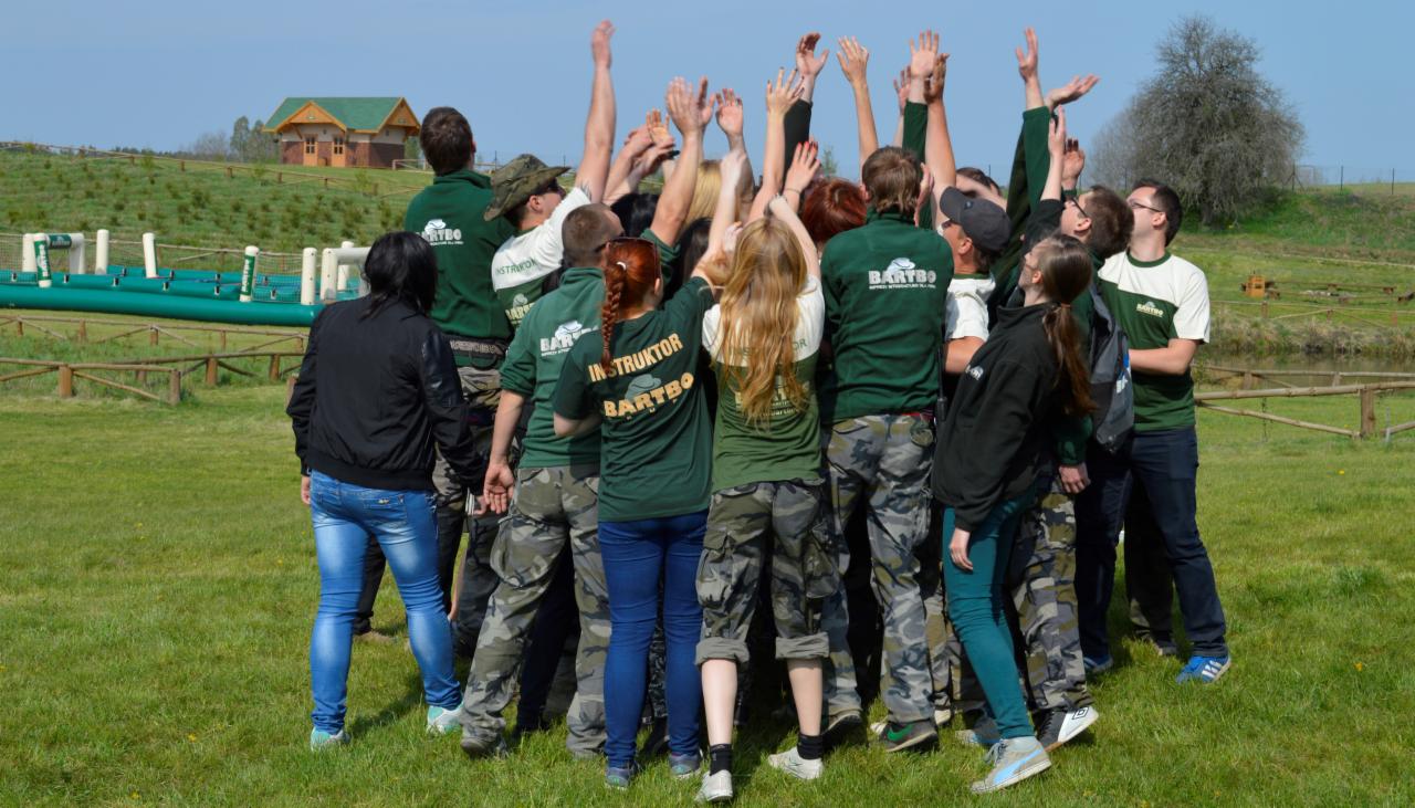 Grupa osób, w tym instruktorzy z napisem 'INSTRUKTOR BARTBO', stojąca na trawie z podniesionymi rękami, prawdopodobnie podczas integracji lub zabawy na świeżym powietrzu, w tle widoczny tor...