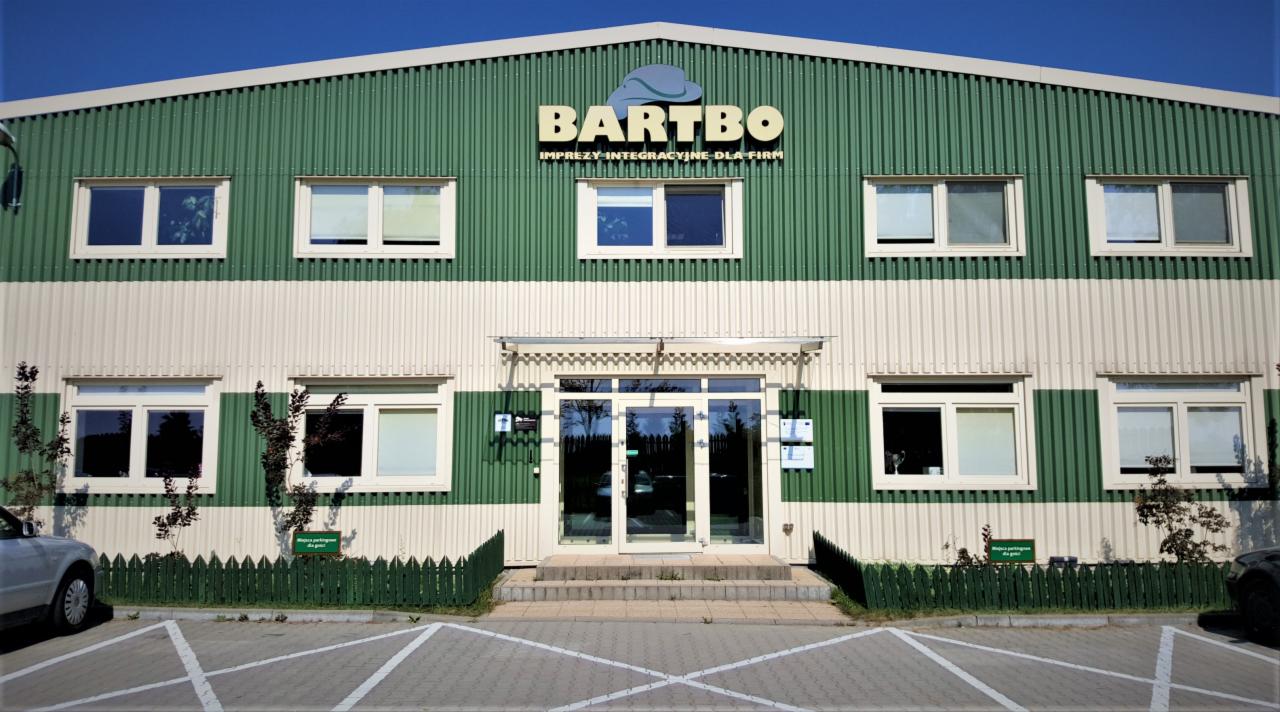 Dwukondygnacyjny budynek o zielono-białej fasadzie z napisem BARTBO i hasłem 'Imprezy integracyjne dla firm' nad wejściem, otoczony niskim płotkiem i parkingiem.