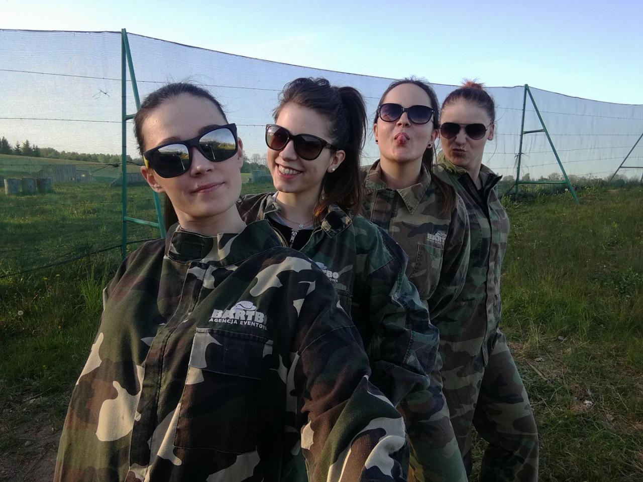 Cztery uśmiechnięte kobiety w okularach przeciwsłonecznych i kombinezonach moro, pozujące na tle pola paintballowego z siatką ochronną, z logo 'BARTB Agencja Eventowa' na kombinezonach.
