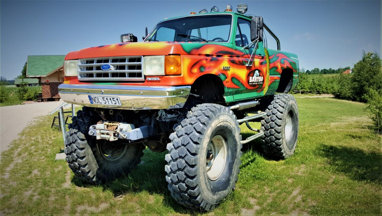 Monster truck z jaskrawym malowaniem w odcieniach zieleni, pomarańczy i żółci, stojący na trawiastym terenie z budynkiem w tle.