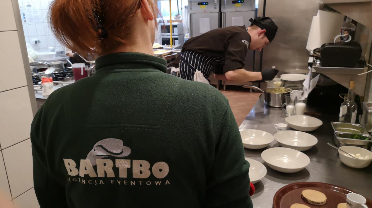 Kucharz w czarnym uniformie i rękawiczkach przygotowuje posiłek w profesjonalnej kuchni, widok zza pleców osoby w zielonej bluzie z logo 'BARTBO Agencja Eventowa'.