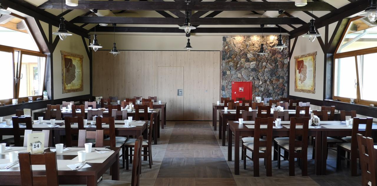 Sala restauracyjna z nakrytymi stołami, drewnianymi krzesłami i dekoracyjną ścianą z kamienia; widoczne menu na stołach.