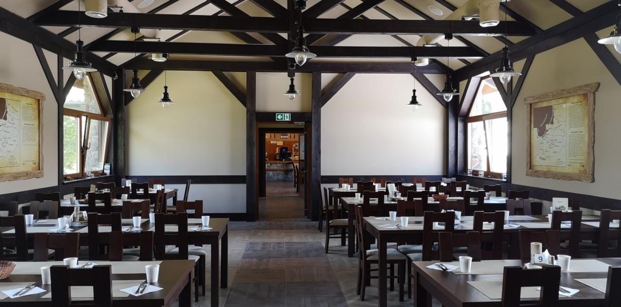 Sala restauracyjna z drewnianą konstrukcją dachu, stołami nakrytymi białymi obrusami i ustawionymi krzesłami, przygotowana na przyjęcie gości, z widoczną tablicą informacyjną na ścianie...