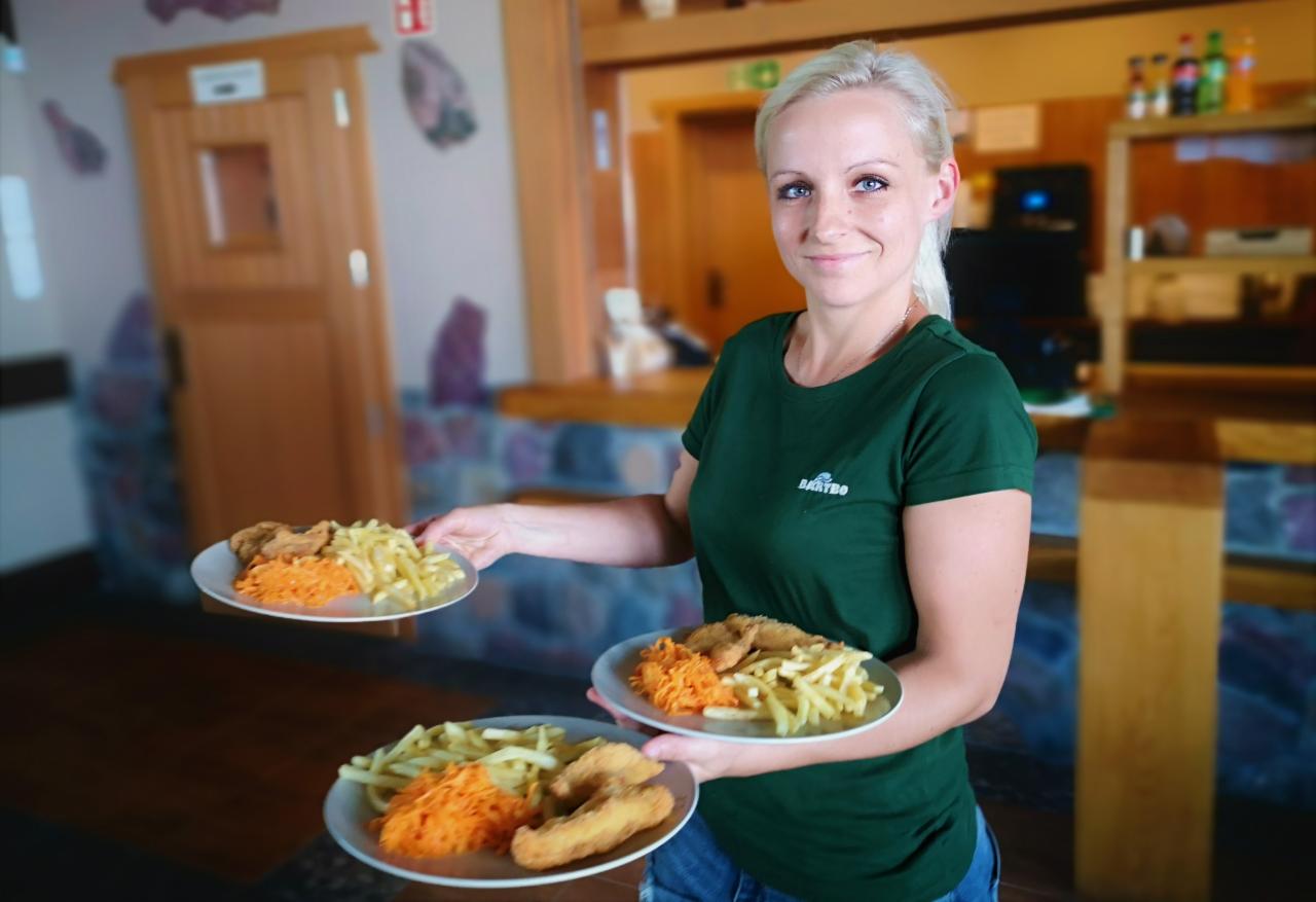 Uśmiechnięta kelnerka w zielonej koszulce z logo firmy trzyma trzy talerze z zestawem obiadowym: smażona ryba, frytki i surówka z marchewki.
