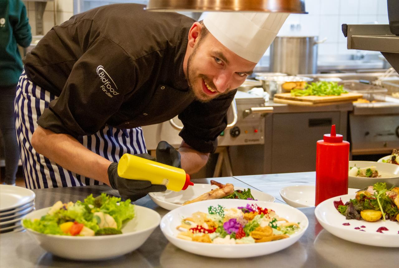 Uśmiechnięty kucharz w czarnym uniformie z logo Bidfood Farutex i czepku, w czarnych rękawiczkach, dekoruje talerze z sałatkami i kwiatami jadalnymi w profesjonalnej kuchni, używając żółtej butelki...