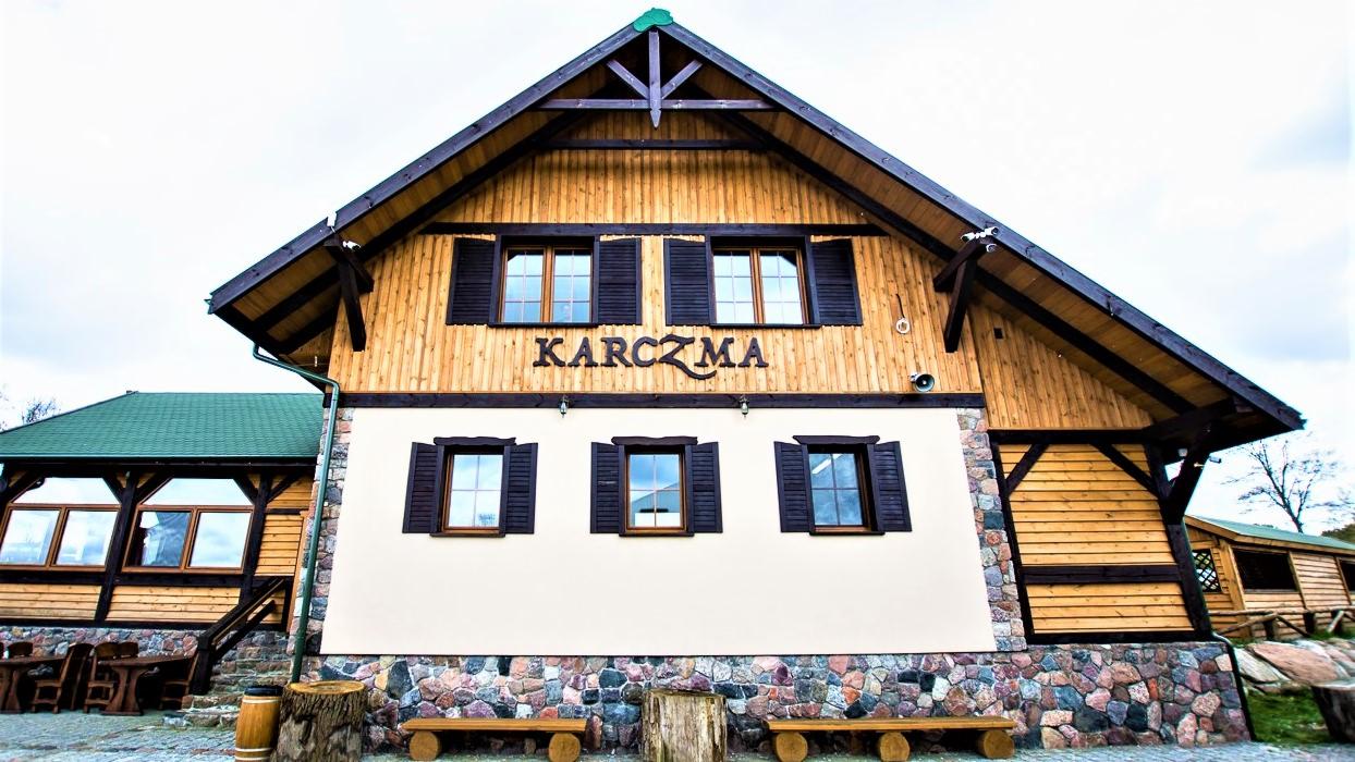 Fasada drewnianej karczmy z napisem 'KARCZMA' nad oknami, kamienna podmurówka, drewniane ławki przed wejściem.