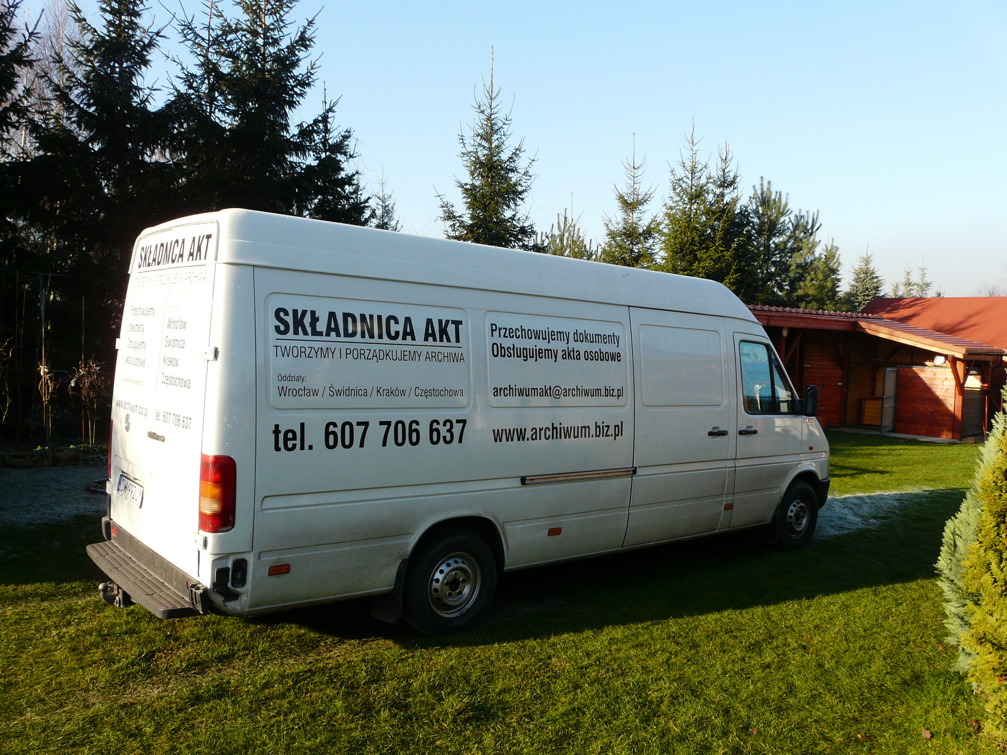 Biały van z napisem 'Składnica Akt' na tle zielonego trawnika i drzew iglastych. Widoczny numer telefonu i adres strony internetowej firmy.