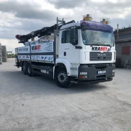 Ciężarówka MAN z dźwigiem marki Krabet, załadowana materiałami budowlanymi, stojąca na betonowym placu. W tle widoczny budynek przemysłowy i stosy zapakowanych elementów.