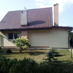 Jasna elewacja domu jednorodzinnego z brązowym dachem, widoczny komin, okno i fragment białego busa na podjeździe, otoczenie to trawnik i krzewy.